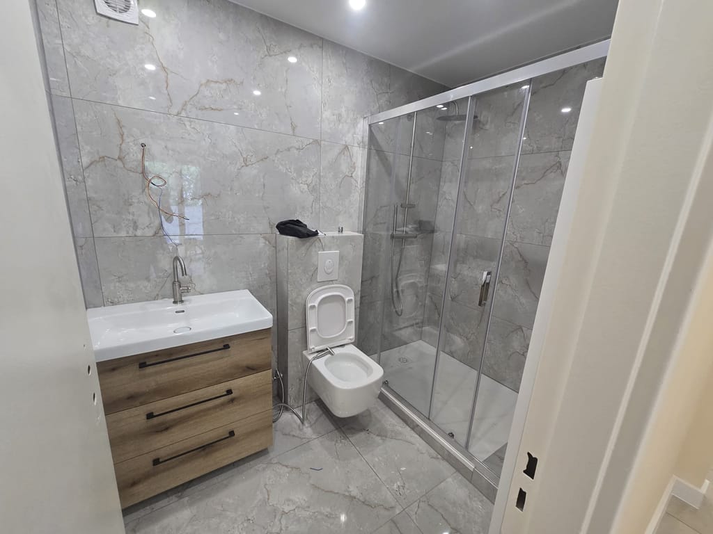 Salle de bains terminée : carrelage gris veiné au sol et sur les murs, douche avec receveur extra-plat fermée par une paroi coulissante, WC suspendu, meuble vasque en bois surmonté d'un plan blanc et spots encastrés au plafond.